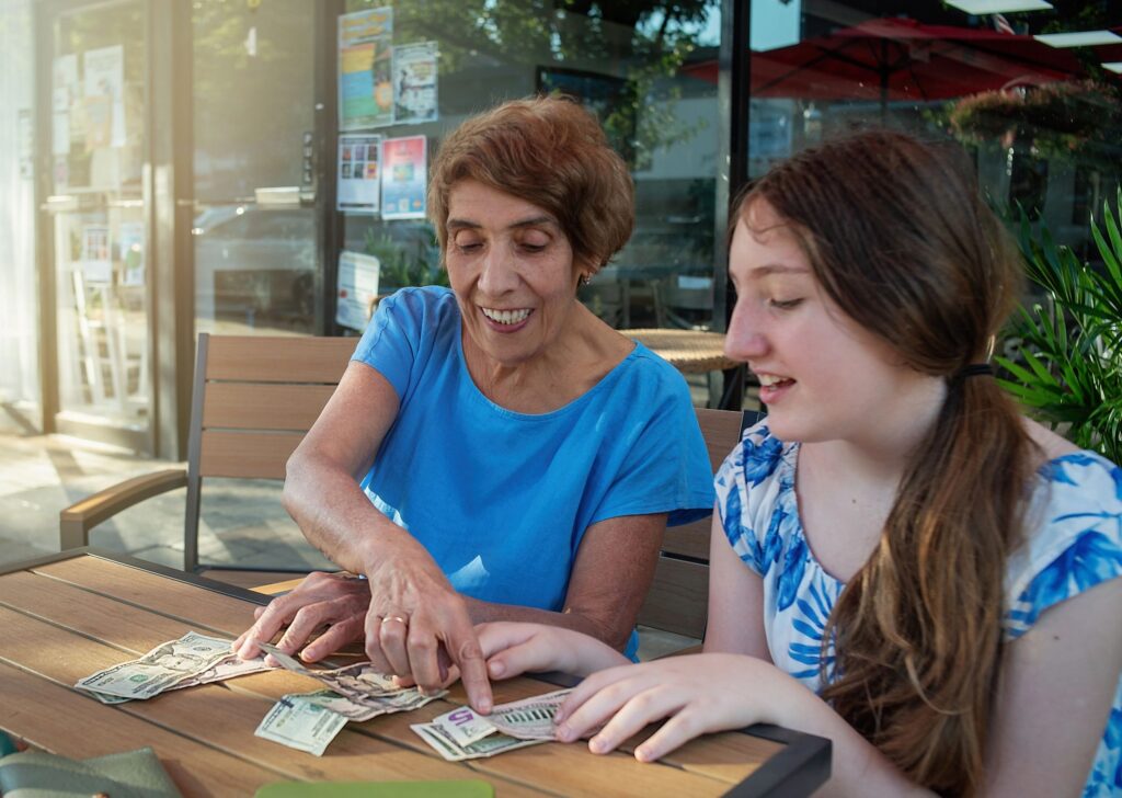 Caucasian senior grandmother and teenage granddaughter actively counting US dollar bills, discussing value of money at a sunny outdoor cafe table. Financial education.